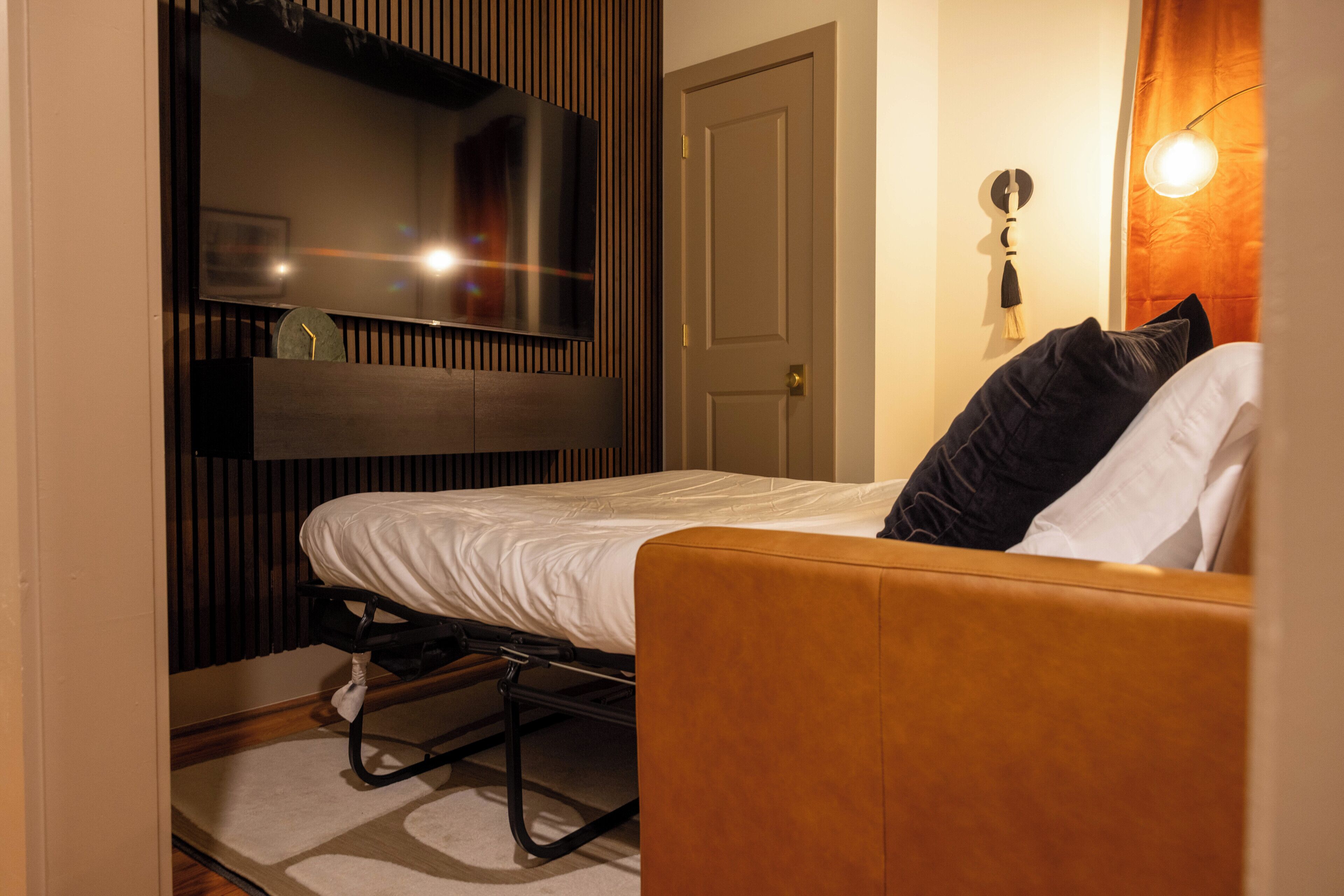 Une chambre à coucher comprenant un lit, un téléviseur fixé au mur, une porte et un luminaire fixé au mur.