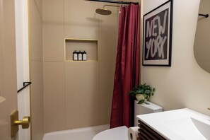 Une salle de bain moderne avec une douche, des toilettes et un lavabo. On y trouve une étagère avec des articles de toilette et un tableau encadré accroché au mur.