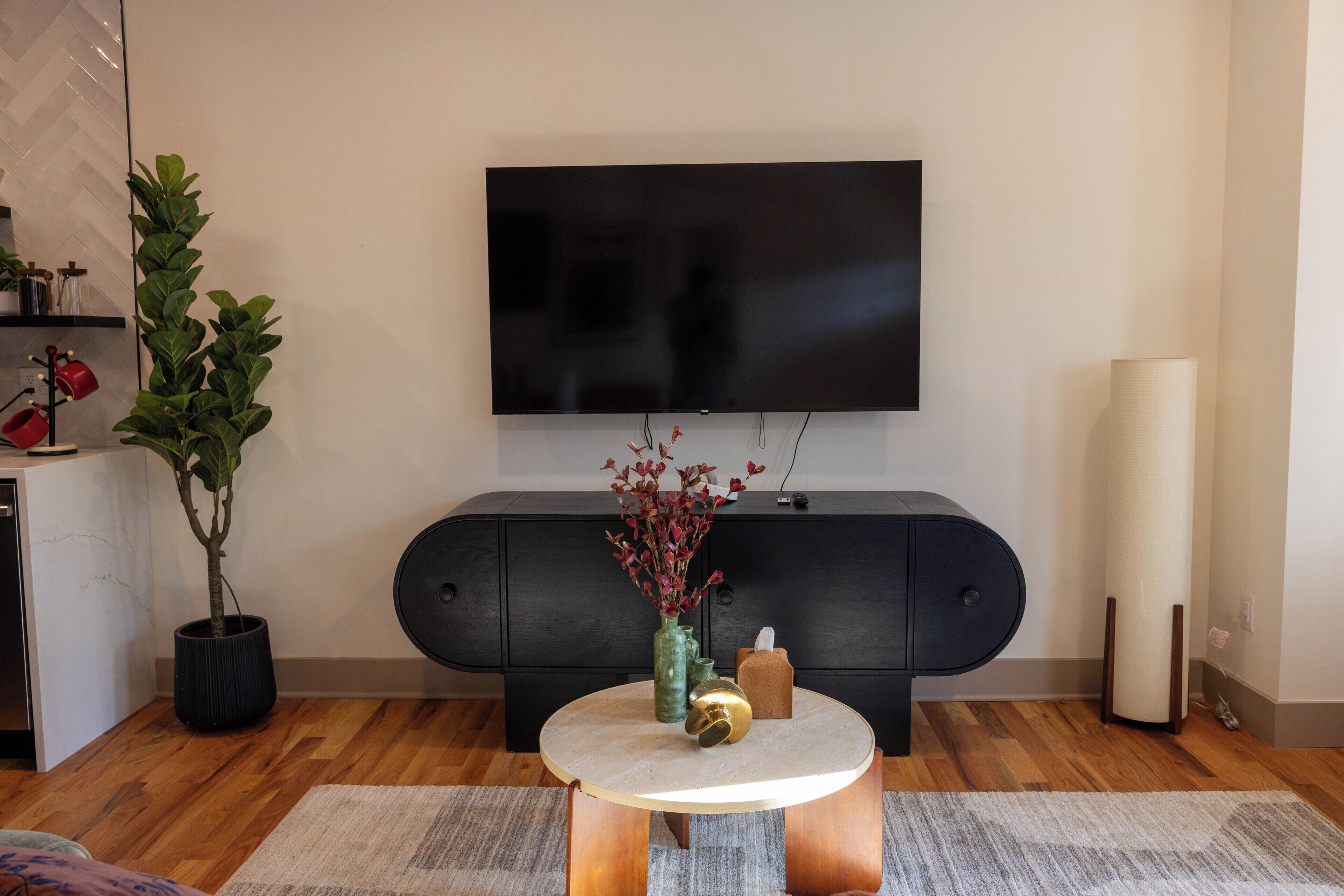 A modern living room with a flat-screen TV mounted on the wall, a dark-colored cabinet, a round coffee table with a vase of flowers, and a potted plant.