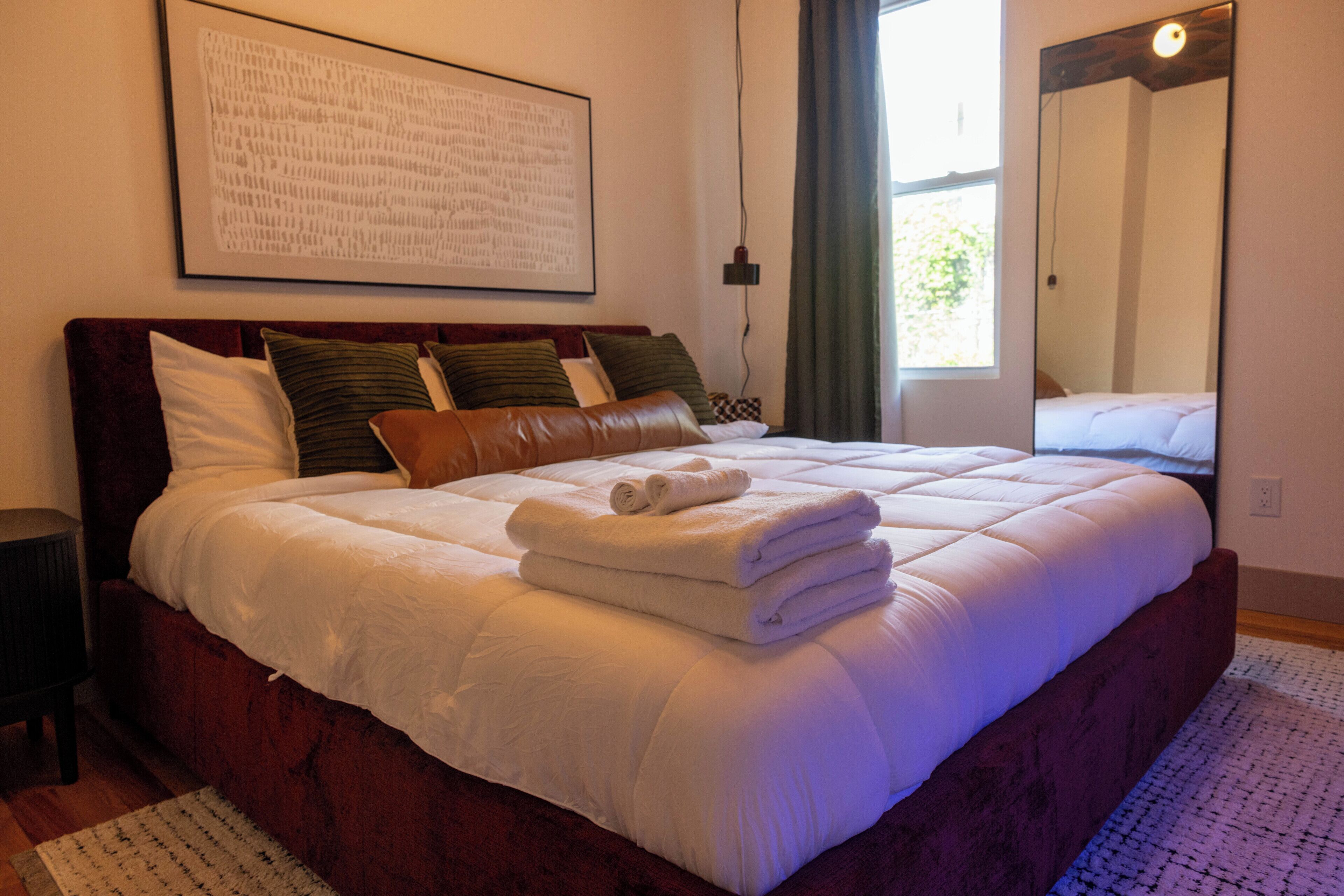 Een netjes opgemaakt bed met kussens en een opgevouwen handdoek, een groot gekaderd kunstwerk aan de muur, en een raam met gordijnen.