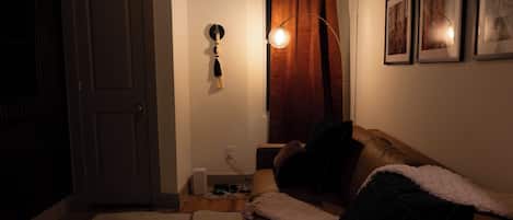 A dimly lit room with a closed door, a standing lamp, a couch with a blanket, and a wall-mounted picture frame.