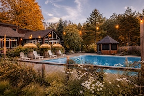 A swimming pool with lounge chairs and umbrellas, surrounded by a wooden fence and trees.
