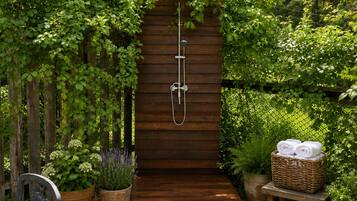 Une douche extérieure en bois, entourée d’une végétation luxuriante, avec un panier en osier rempli de serviettes posé sur un tabouret à proximité.