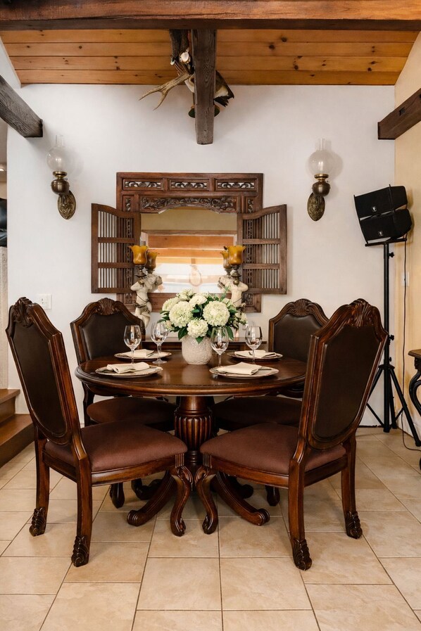 A dining room with a round table, six chairs, a vase of flowers, and a mirror with shutters.