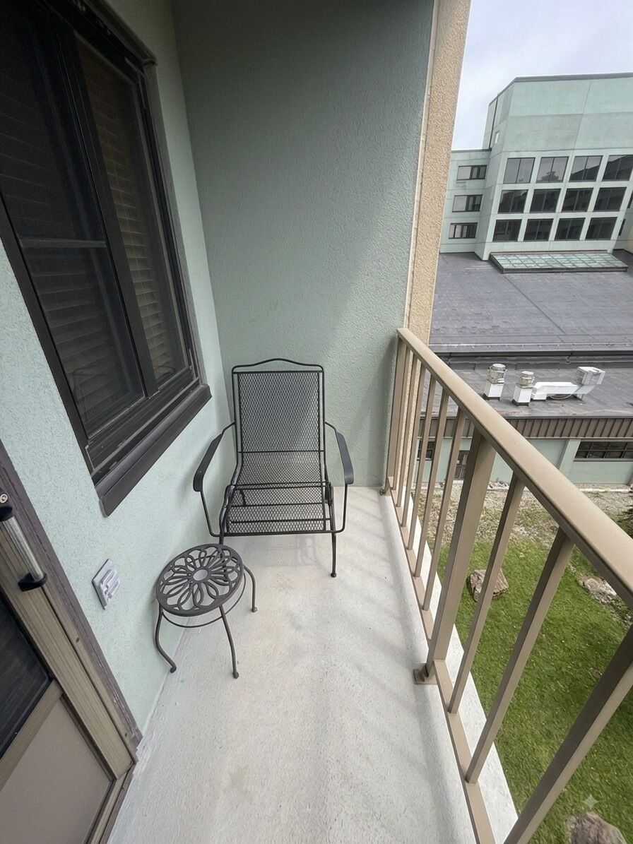 A balcony with a metal chair and a small table, a window with blinds, and a building in the background.
