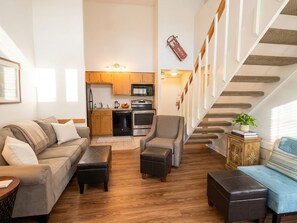 Ein modernes Wohnzimmer mit Sofa, Sesseln und Hocker, im Hintergrund eine Küche und eine Treppe mit Geländer.