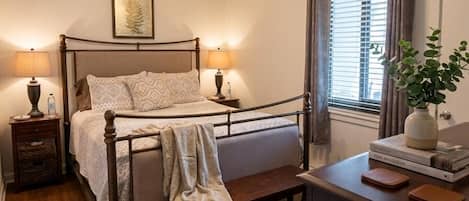 Een slaapkamer met een bed, nachtlampjes, een plafondventilator, een raam met gordijnen, een nachtkastje en een bureau met een plant en boeken.