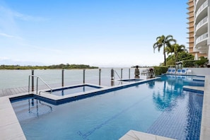 A swimming pool with a view of the water body, a palm tree, and a building with balconies.