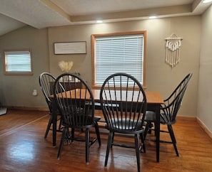 A dining area with a wooden table and four chairs, a window with blinds, and a wall-mounted macrame decoration.