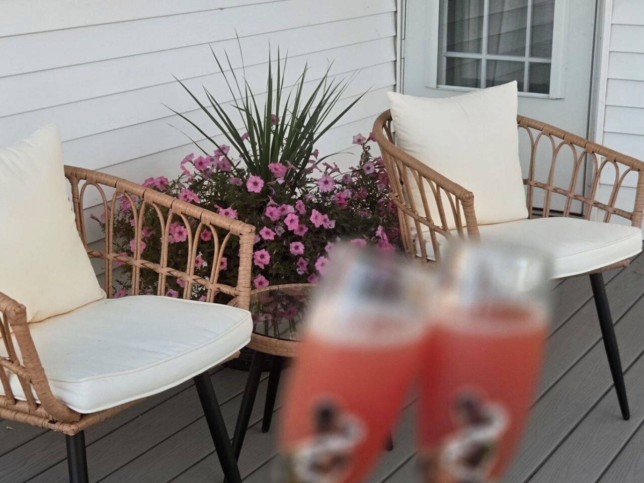 A wicker chair and table set on a porch with a potted plant and two glasses of red beverage.