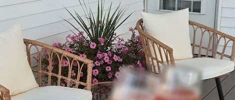 A wicker chair and table set on a porch with a potted plant and two glasses of red beverage.