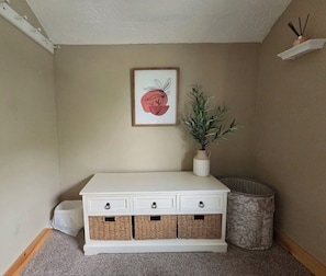 A white storage bench with wicker baskets, a framed artwork, and a potted plant against a beige wall.