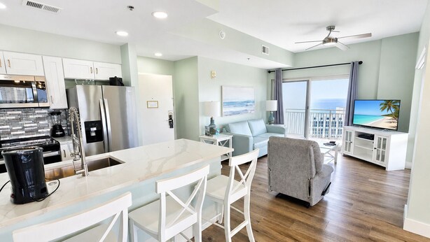 A modern kitchen with a marble countertop and white bar stools, a dining area with a white table and chairs, and a living room with a sofa and a television displaying a beach scene. - 10th-Floor Luau Condo | Fabulous South-Facing Gulf Views & Beach Access (Miramar Beach)