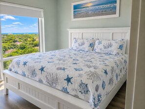 Un dormitorio con un ventanal grande, una cama con colcha de diseño de estrella de mar y coral, y un cuadro enmarcado con una escena de playa en la pared.