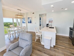 A modern kitchen with a white island, wooden flooring, and a dining area with white chairs and a gray sofa. - 10th-Floor Luau Condo | Fabulous South-Facing Gulf Views & Beach Access (Miramar Beach)