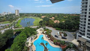 A view from a balcony overlooking a swimming pool, a golf course, and a residential area with a body of water and a tall building in the distance. - 10th-Floor Luau Condo | Fabulous South-Facing Gulf Views & Beach Access (Miramar Beach)