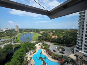 Vista desde un balcón sobre una piscina, un campo de golf y una zona residencial con una masa de agua y un edificio alto a lo lejos.