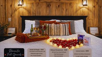 Un dormitorio acogedor con cabecera de madera, una mesita de noche con una botella, vasos y una bandeja de pasteles. Hay velas y pétalos de rosa sobre la cama, y un cuadro en la pared.