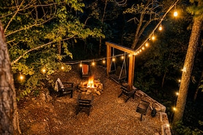 Un'area salotto esterna con un braciere, sedie di legno e luci a stringa.