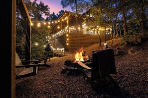 A wooden deck with a fire pit, surrounded by chairs and a swing set, under string lights. - Private & Romantic | Hot Tub, ATV Trails access (Broken Bow)