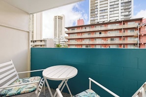A balcony with a white table and chairs, a teal wall, and a view of a multi-story building with balconies. - Steps to Waikiki Beach | Prime Beach Location! (Honolulu)