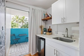 A compact kitchen with a dishwasher, two sinks, and a window with a view of palm trees. - Steps to Waikiki Beach | Prime Beach Location! (Honolulu)