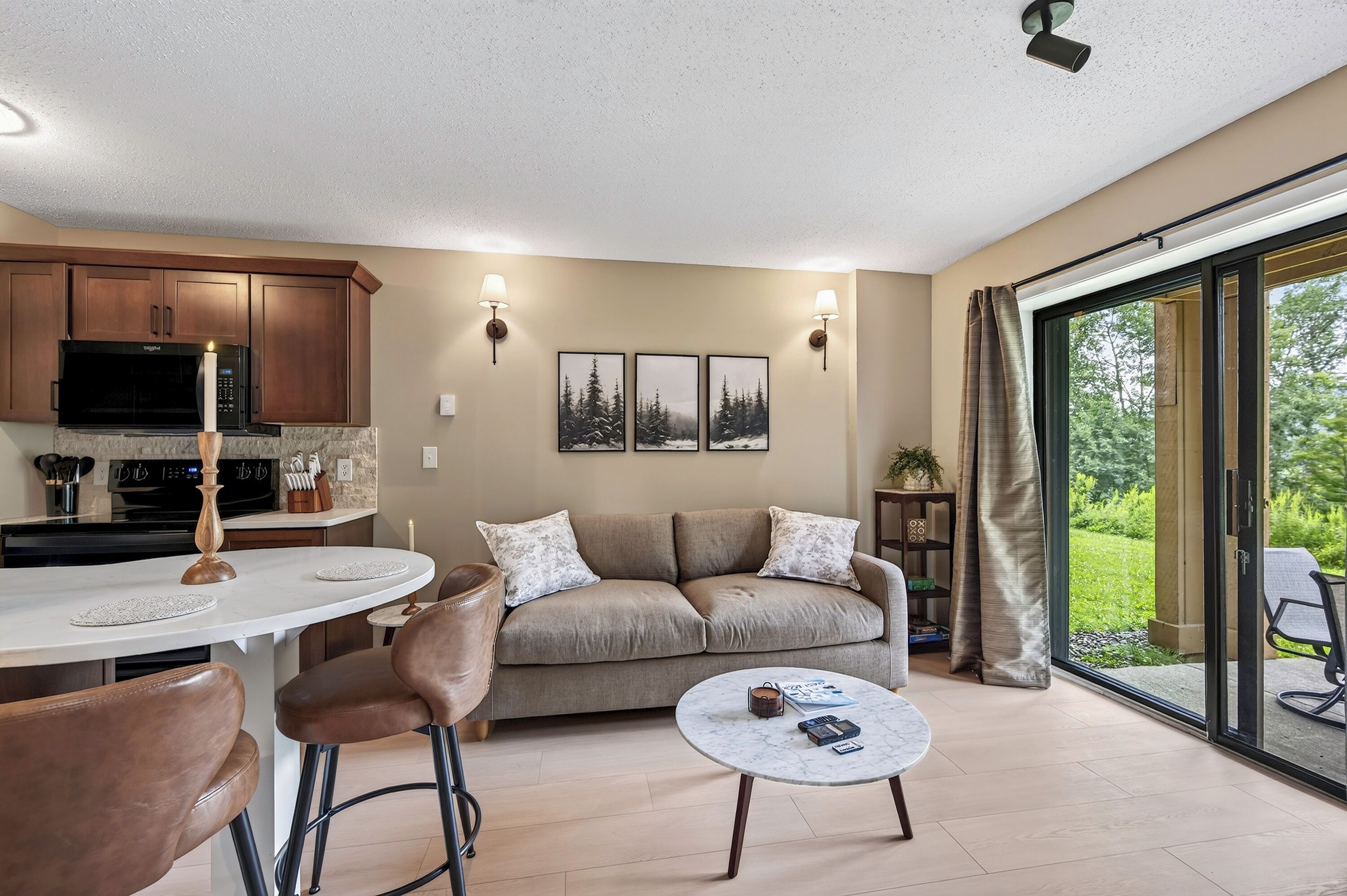 A modern kitchen with a microwave, stove, and cabinets, a dining area with a table and chairs, a sofa with cushions, and a large window with a view of greenery.