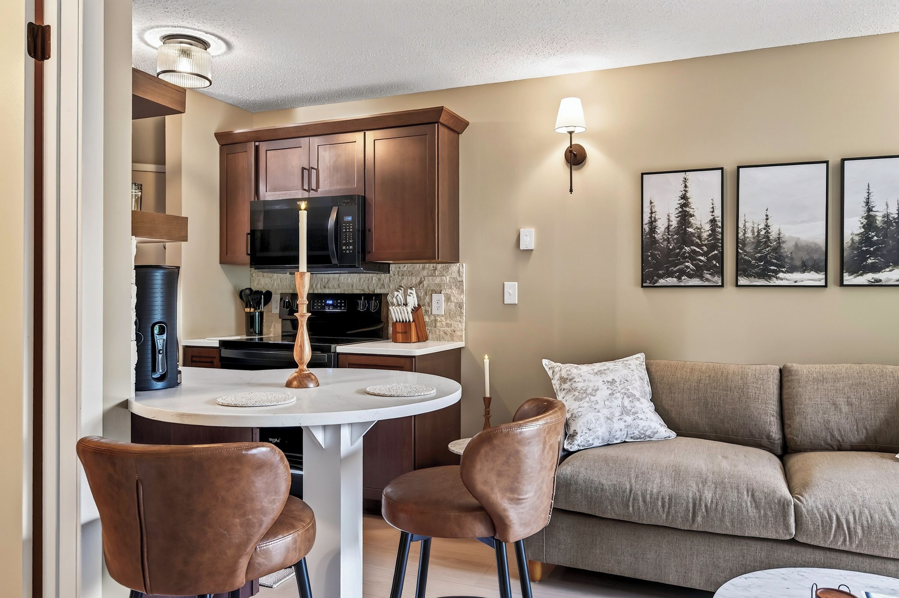 A modern kitchen with dark wooden cabinets, a black microwave, and a gas stove. There is a round dining table with two chairs and a candle on it. A beige sofa is in the living area with a mounted picture frame.