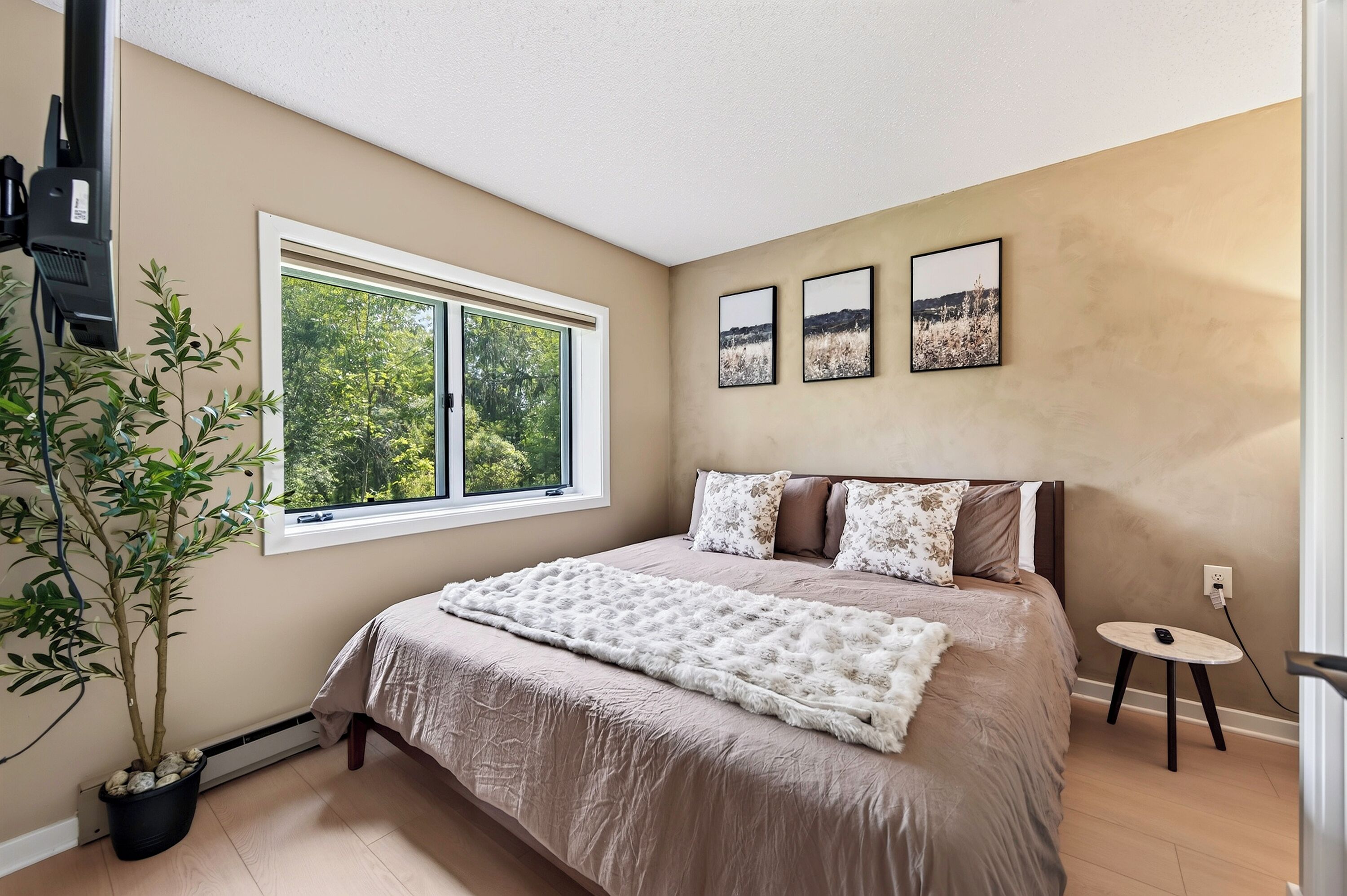 A bedroom with a large window, a bed with a fluffy white throw, and two framed pictures on the wall.
