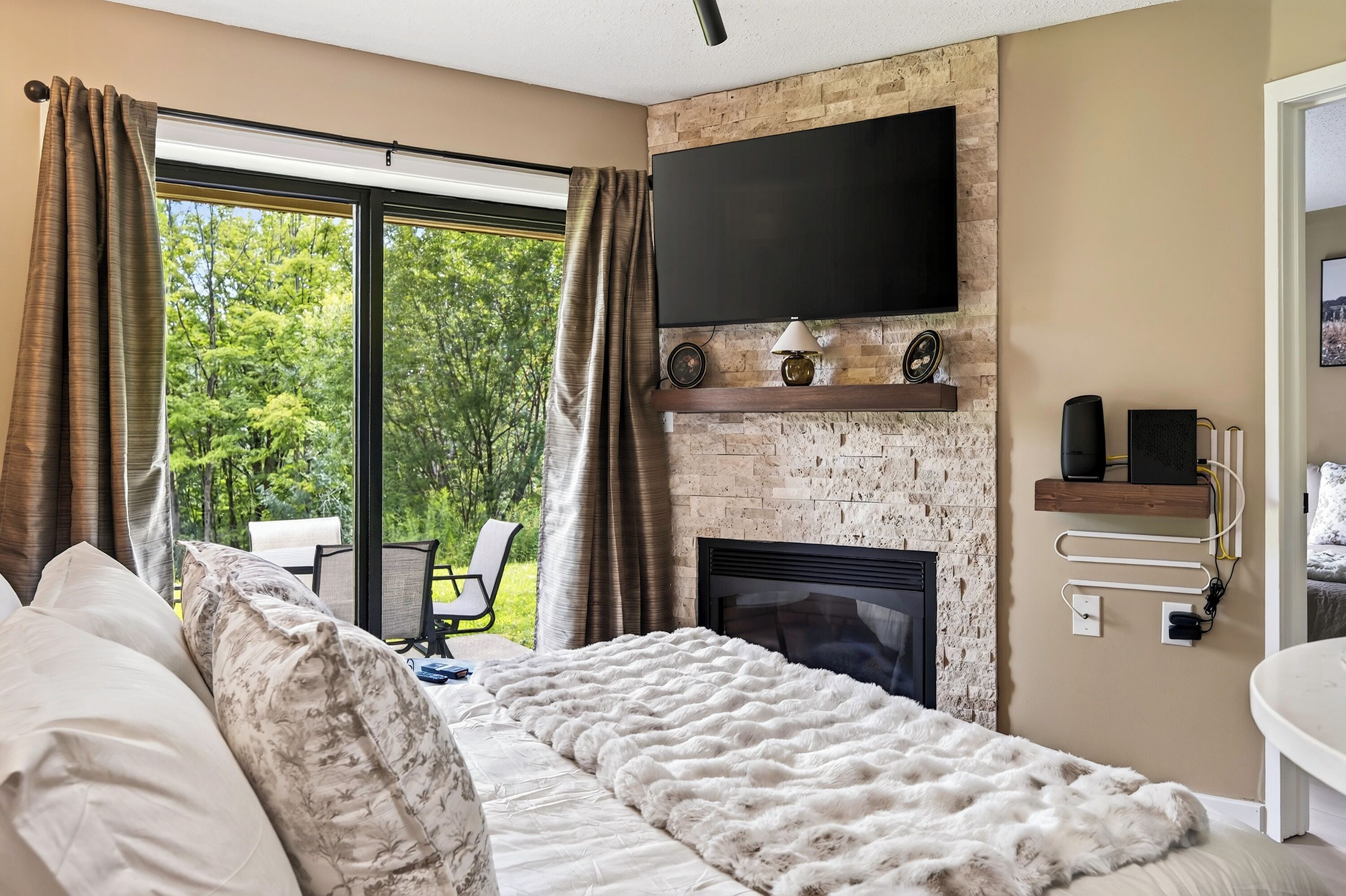 A bedroom with a fireplace, a large window with curtains, a bed with a fluffy white blanket, and a TV mounted on the wall.