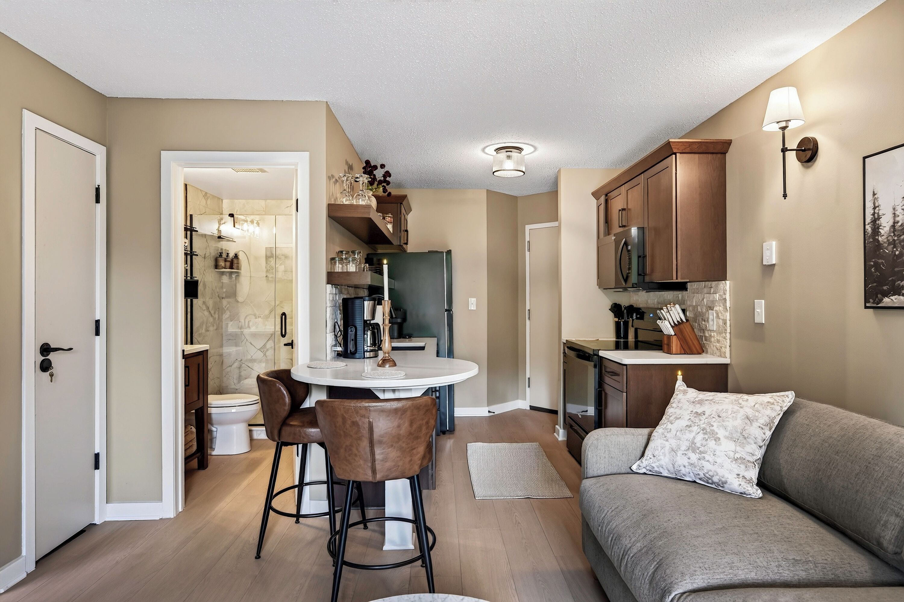 A compact living space with a kitchenette, a dining area with a round table and chairs, a sofa with a cushion, and a shower area.