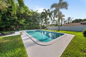 Ein rechteckiger Pool mit Betonrand, umgeben von Gras und verschiedenen tropischen Pflanzen.
