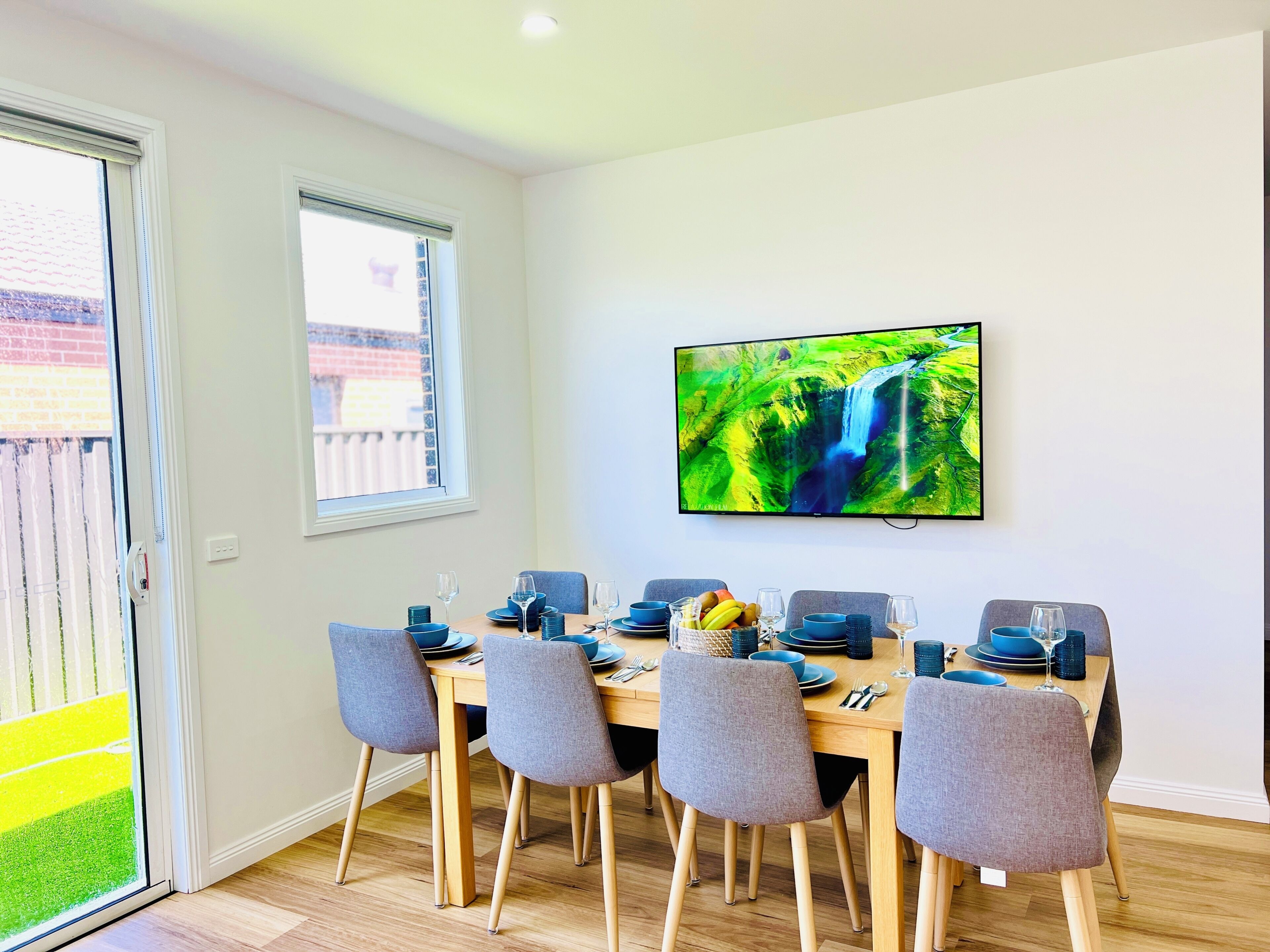Un coin repas avec une table dressée pour six personnes, des chaises, un téléviseur fixé au mur et une porte-fenêtre coulissante donnant sur un espace extérieur.