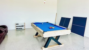 A pool table with balls, a cue stick, and a blue tablecloth in a room with white walls and a speckled floor.