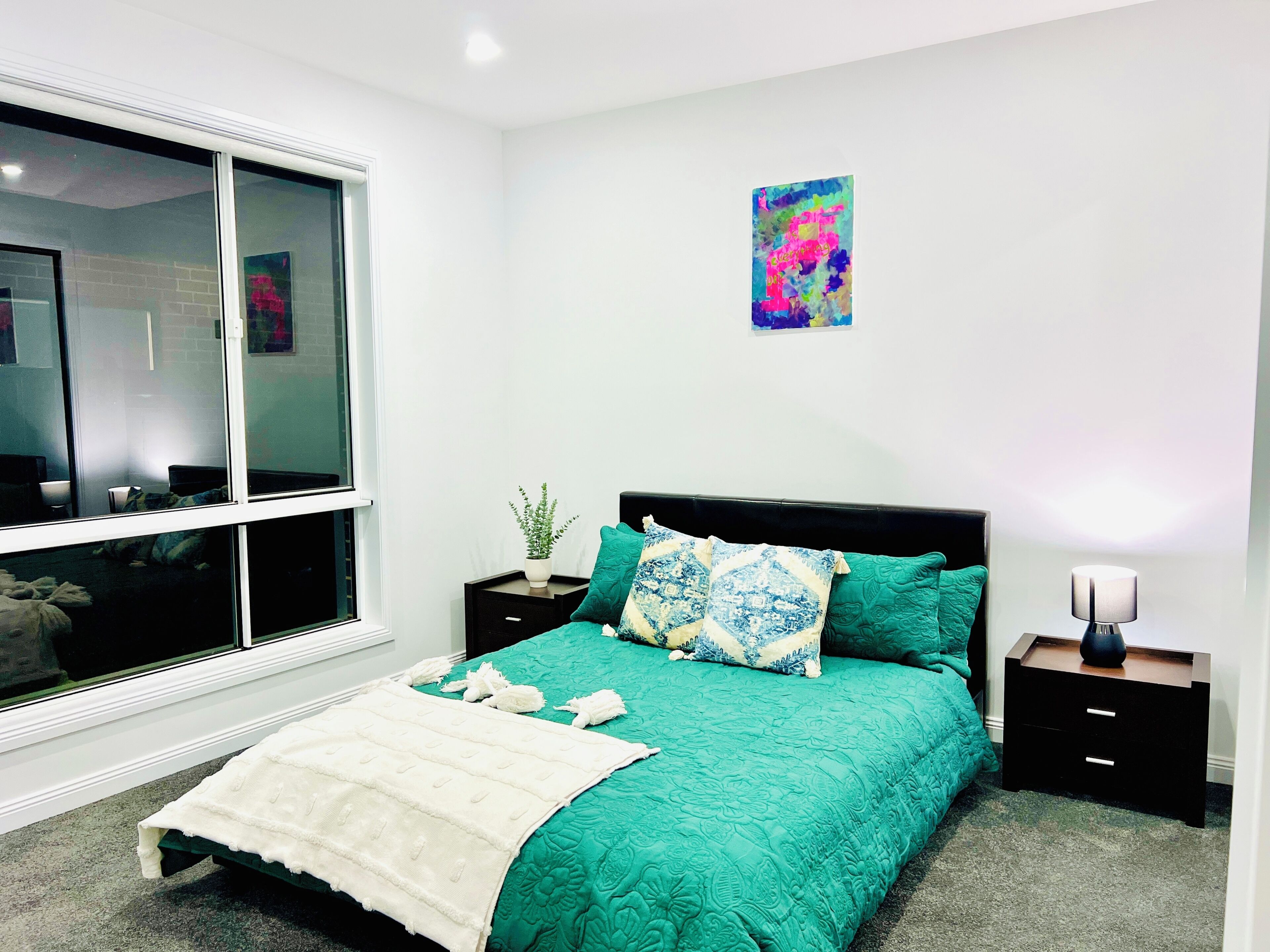 A bedroom with a bed, a bedside table, a nightstand, a window, and a wall-mounted artwork.