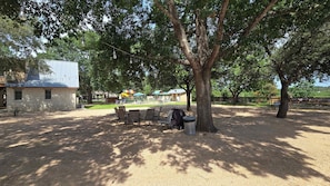 An outdoor seating area with tables and chairs under trees, a barbecue grill, and a building with a stone facade in the background. - Rosemary Gruene Cottage Studio (New Braunfels)
