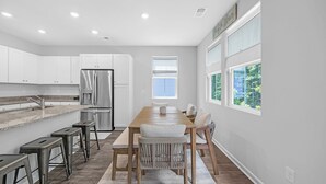 A modern kitchen with white cabinets, stainless steel appliances, and a long wooden dining table with bar stools. - Enjoyable 4-bedroom house with AC in charming Little River (Little River)