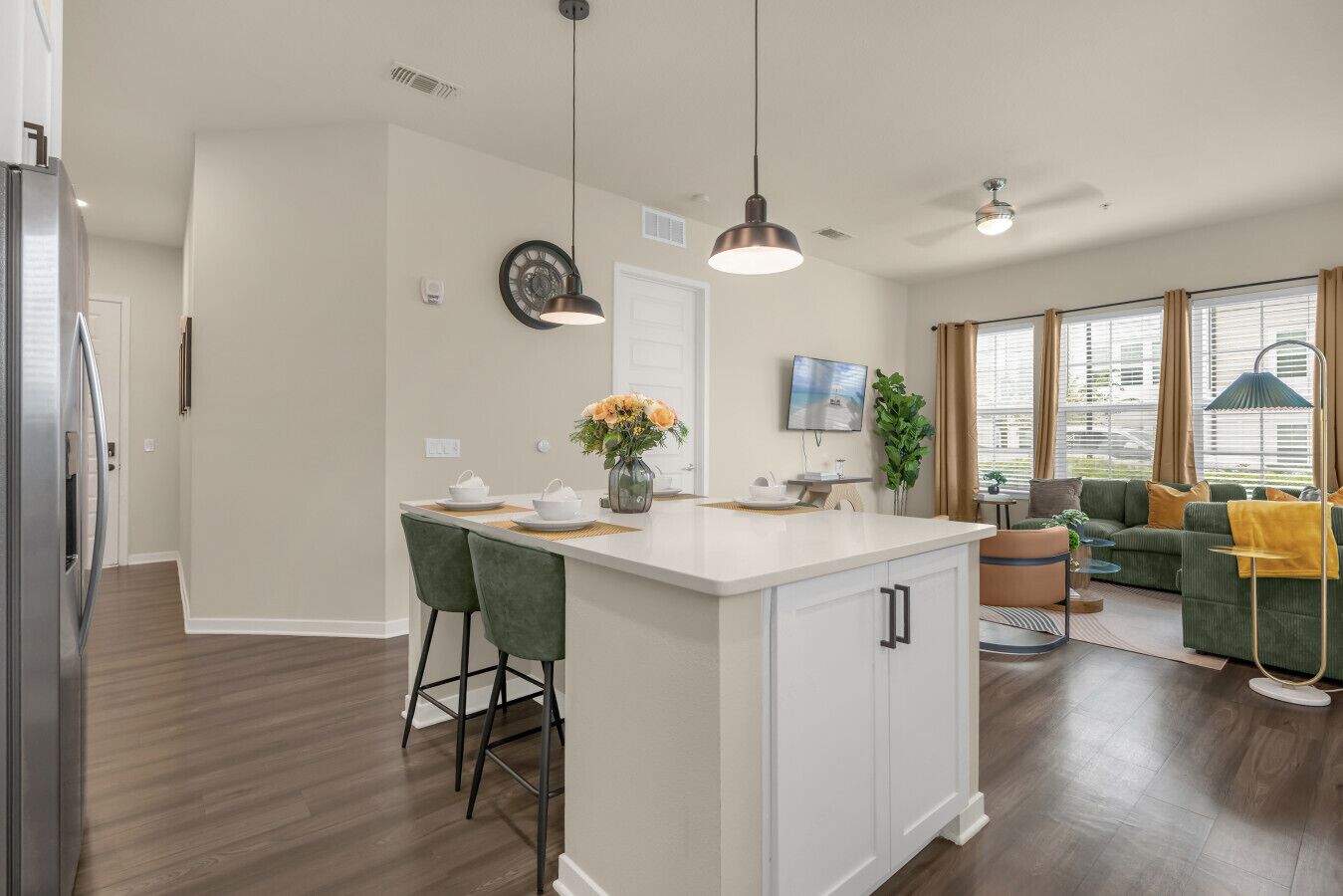 A modern kitchen with a central island, bar stools, pendant lighting, and a dining area with a sofa and a TV.