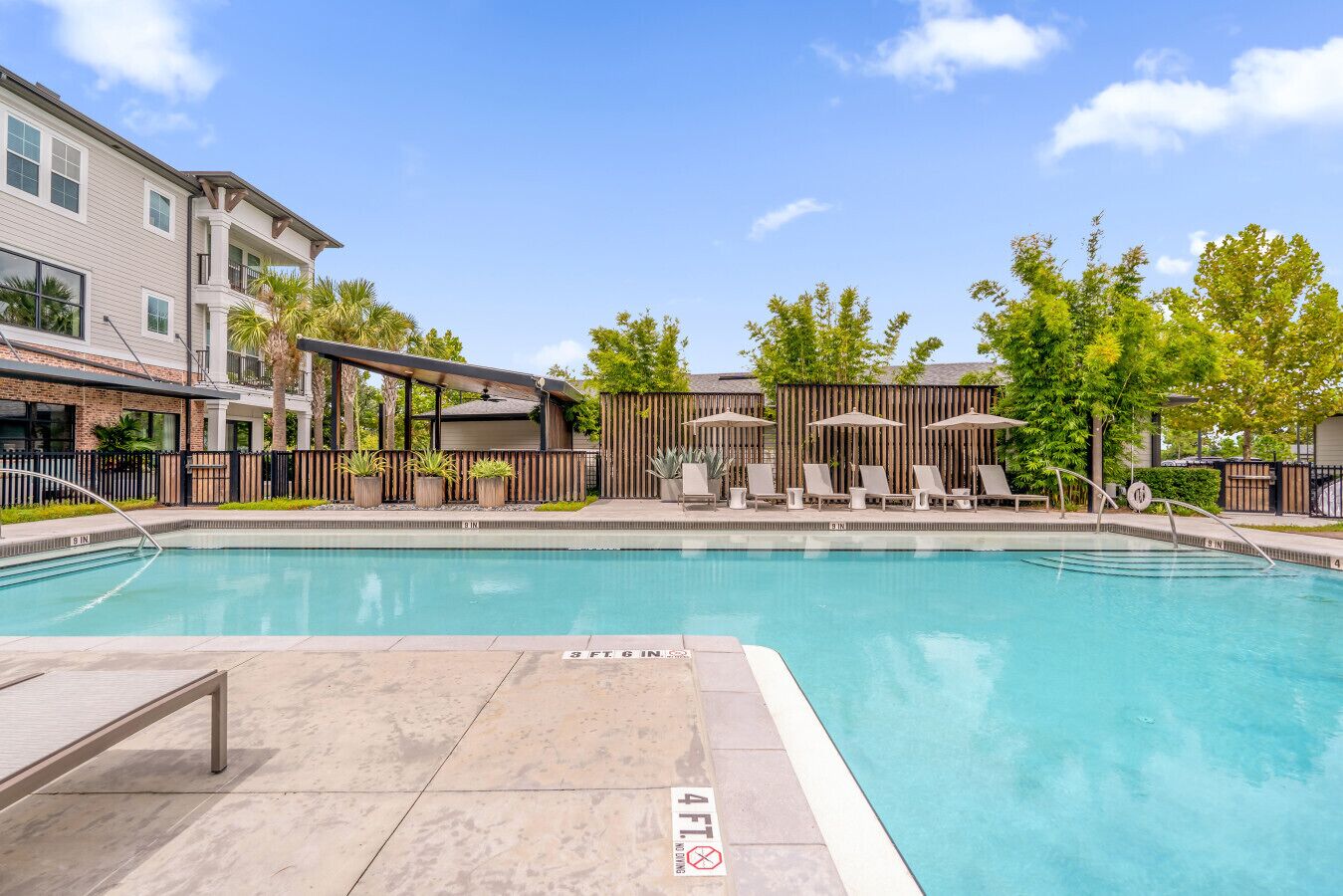 A swimming pool with lounge chairs and umbrellas, surrounded by a fence and trees.