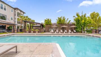 A swimming pool with lounge chairs and umbrellas, surrounded by a fence and trees.