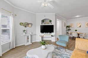 En moderne stue med et fladskærms-tv, en loftventilator, et spiseområde med bord og stole, samt et lille hvidt konsolbord med dekorative genstande.