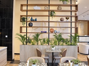 A modern interior with a wooden shelving unit displaying decorative items and plants, a seating area with tables and chairs, and a reception desk in the background. - Citadines Sharq Kuwait (Kuwait City)