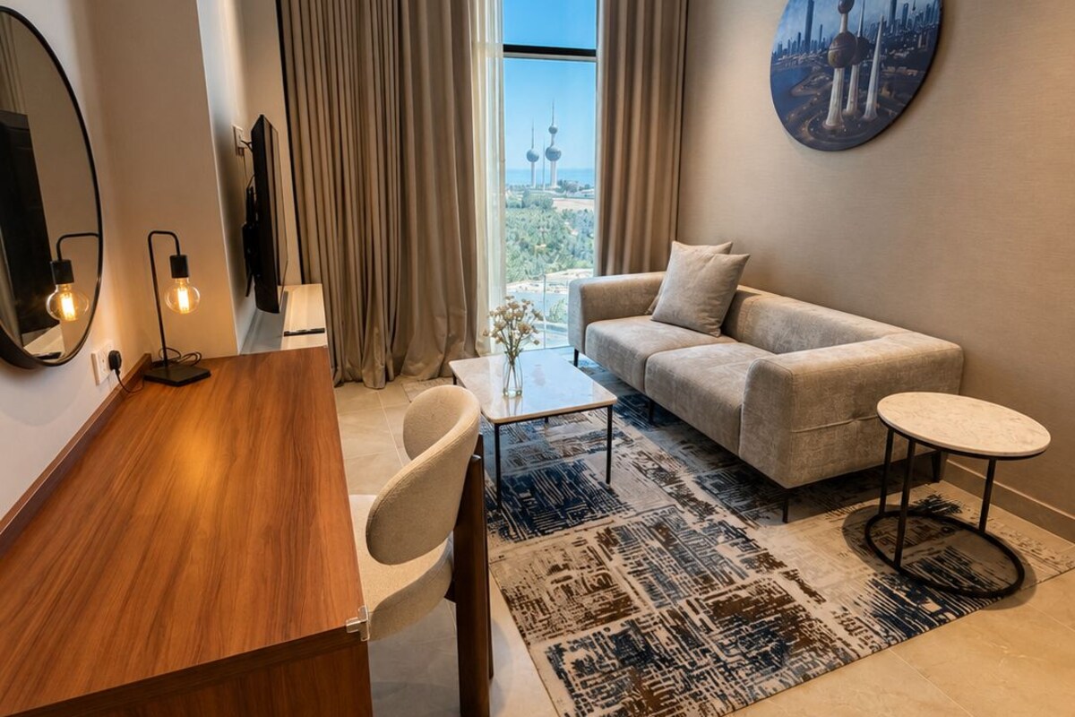 A modern living room with a sofa, coffee table, and a desk with a chair. There is a large window with curtains, a round painting on the wall, and a television on the wall.