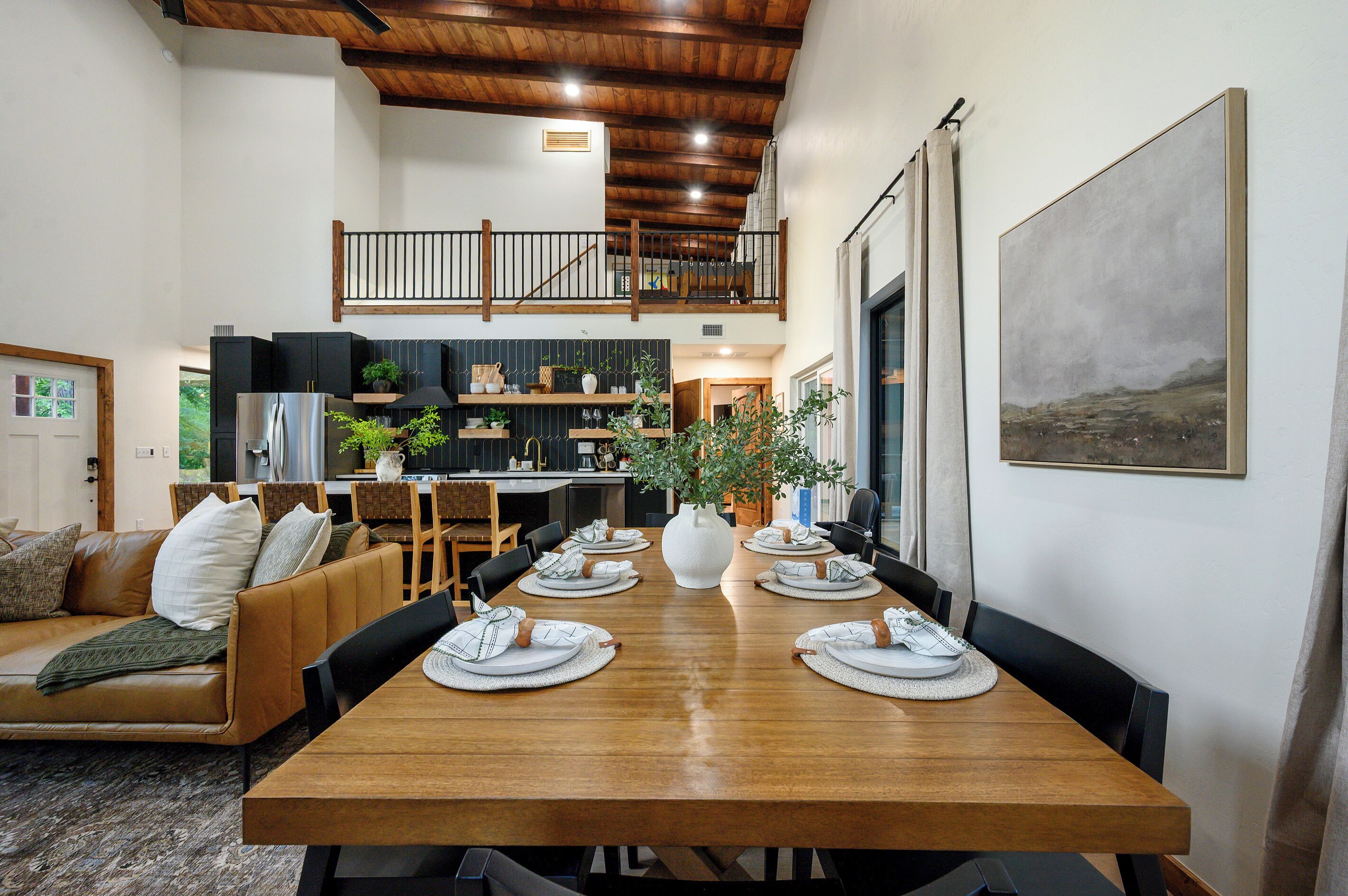 A dining area with a wooden table set for six, a sofa with cushions, and a kitchen in the background.