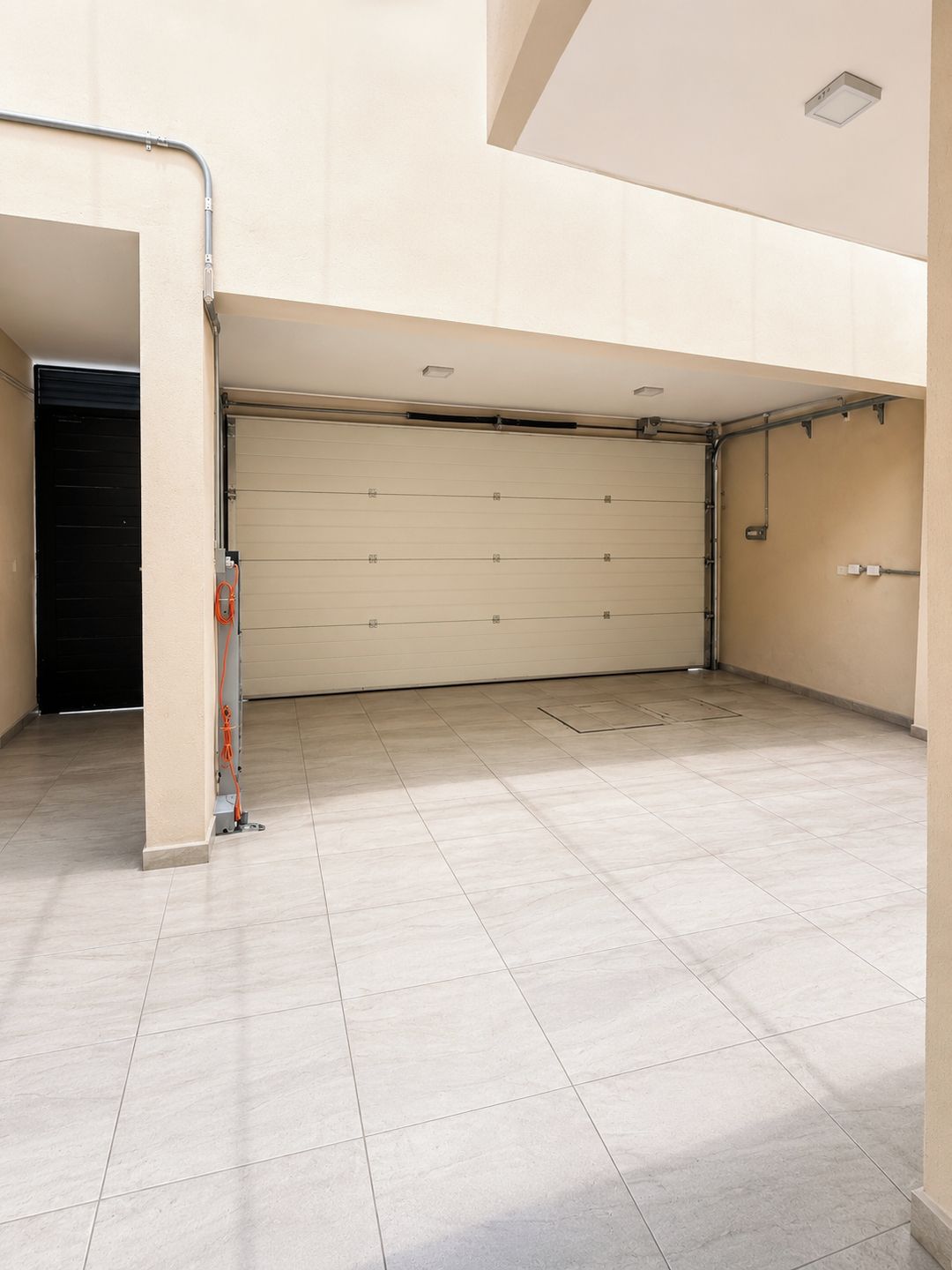 A clean, empty garage with a closed garage door, tiled floor, and visible electrical wiring.