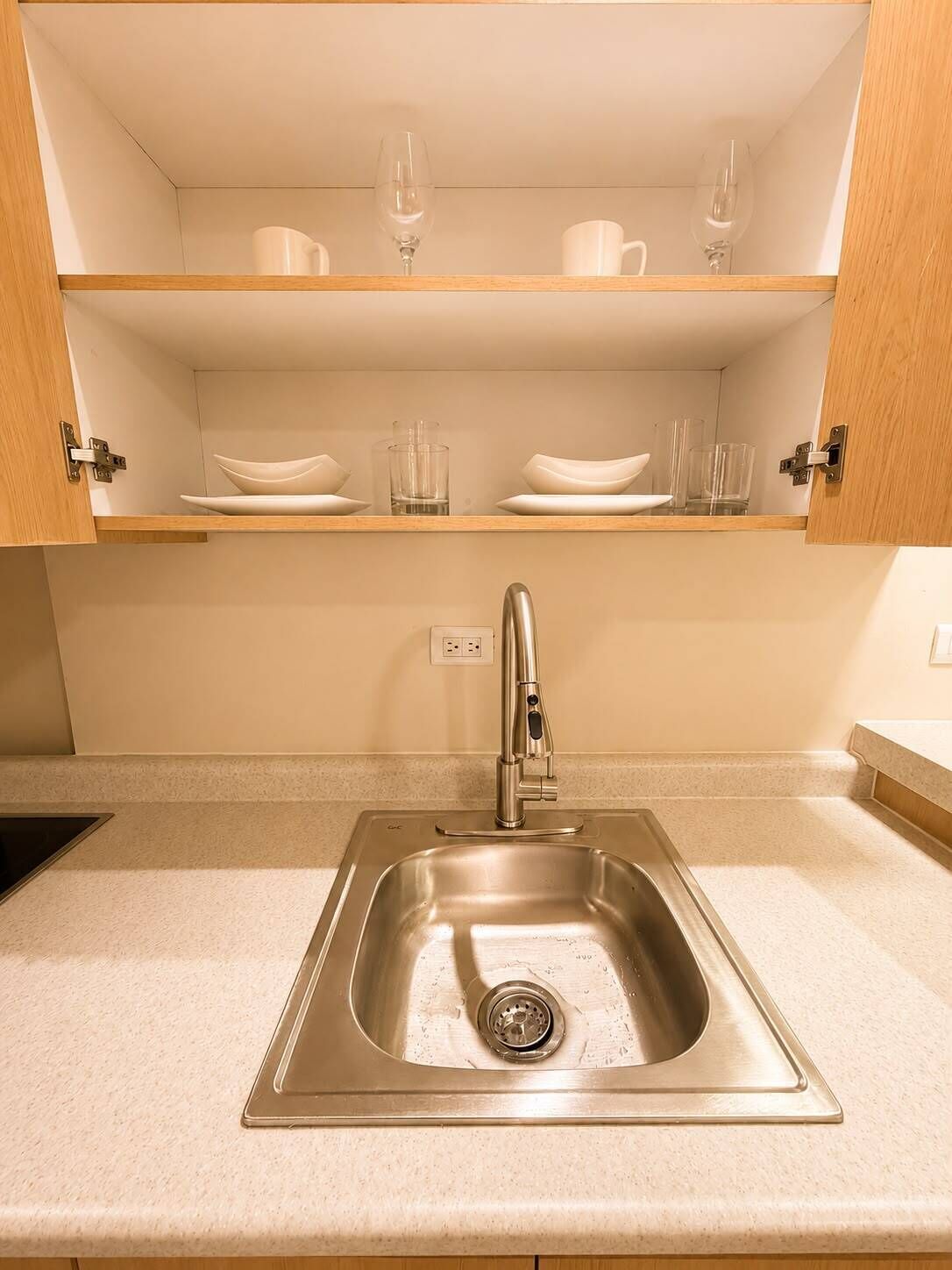A kitchen with a stainless steel sink, wooden cabinets, and shelves with white dishes and glasses.