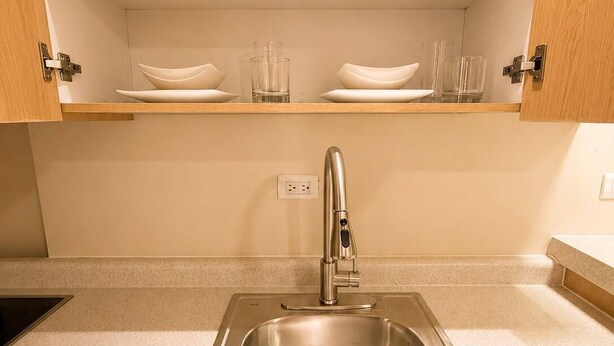 A kitchen with a stainless steel sink, wooden cabinets, and shelves with white dishes and glasses.