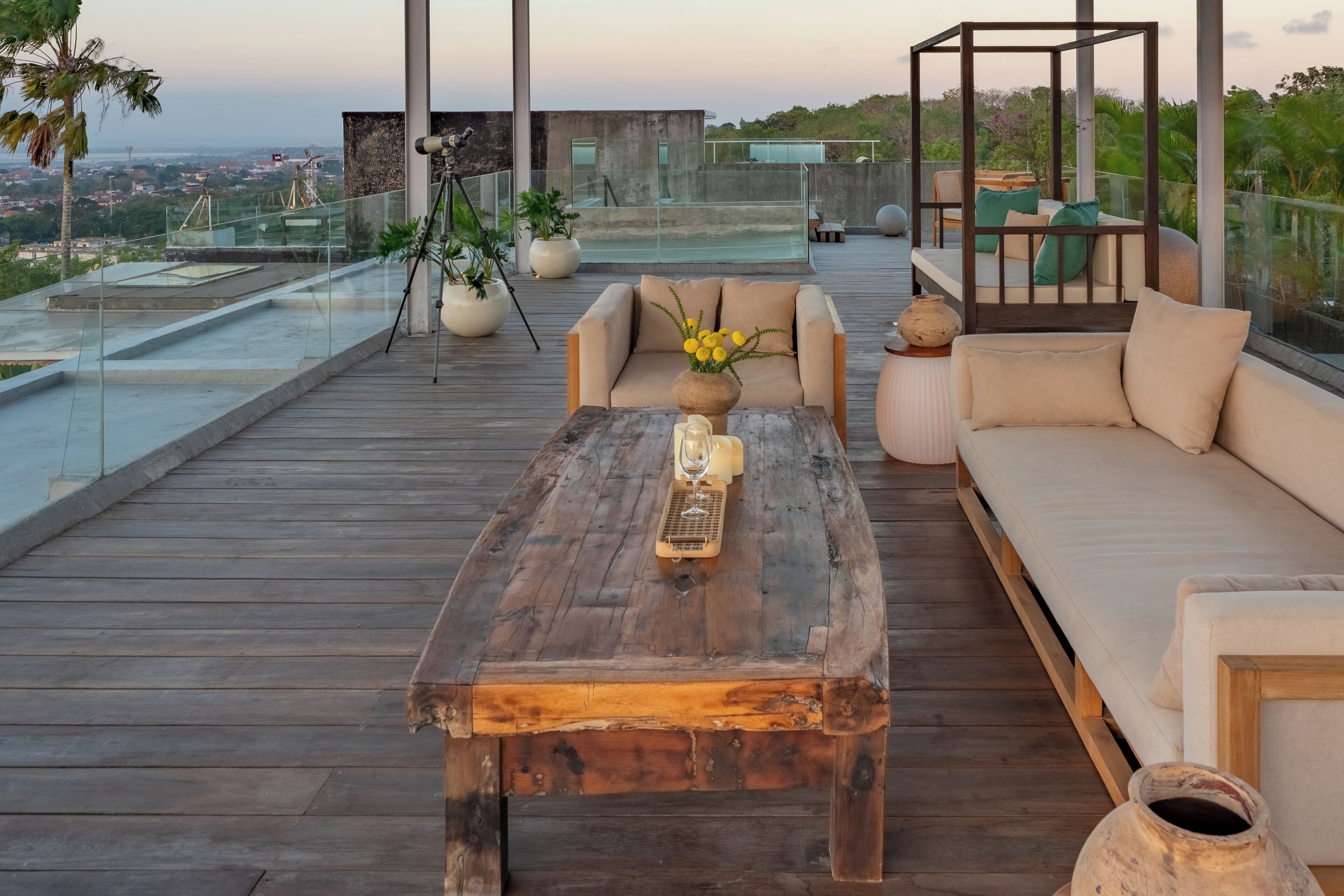 A wooden deck with a sofa, a coffee table, and a glass railing overlooking a cityscape.