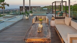 Una terraza de madera con un sofá, una mesa de centro y una barandilla de cristal con vistas a un paisaje urbano.