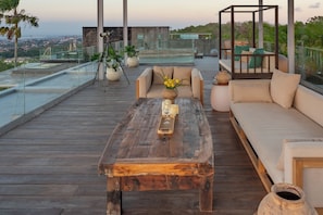 A wooden deck with a sofa, a coffee table, and a glass railing overlooking a cityscape.
